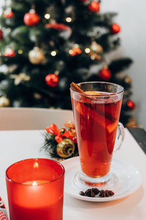 Close of candle and cup warm winter drink for holiday at white table on background decorated Christmas tree. Happy new year concept. selective focus. Mulled wine with cinnamon and orange fruitの写真素材