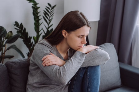 Close up of sad pensive young caucasian woman looking down depressed sitting on sofa in living room at home, thinking about personal problem, break up with boyfriend or divorce. Feeling lonely conceptの写真素材