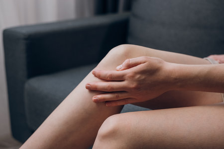 Closeup of unhealthy young woman sitting on sofa and feeling knee pain, doing massage knee at home. Female legs with meniscus injury or chronic joint inflammation. Health care and medical conceptの写真素材