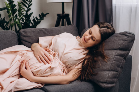 Adorable happy young pregnant woman in dress stroking belly, feeling unconditional love to unborn baby, expressing caress and devotion, enjoying carefree serene weekend lying on cozy couch at homeの写真素材