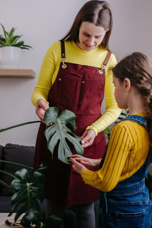 Caring young pregnant mother tells daughter how to take care of house plants at home. Concentrated little girl kid helping mom to care plants. Concept of hobby, schooler leisure and parentingの写真素材