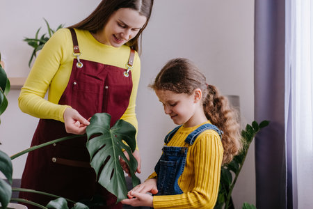 Charming young mother with adorable little daughter cleans indoor plants, takes care green leaves monstera. Gardening, housewife and housework chores concept. Home hobbies, weekend and growing plantsの写真素材