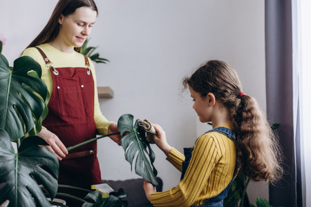 Close up of young mother and little daughter cleans indoor plant, takes care green leaves monstera. Gardening, housewife and housework chores concept. Home hobbies, loving family and growing plantsの写真素材