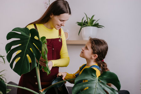 Happy young mother advising pretty little daughter how to water plant in pot at home. Home hobbies, growing plants, gardener or florist work small shop concept. Cute girl kid helping smiling momの写真素材