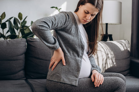 Side view of upset pregnant young female suffering lower back pain sitting on sofa in living room. Sad future mother suffer from spine muscle ache backache. Concept pregnancy, motherhood, healthの写真素材