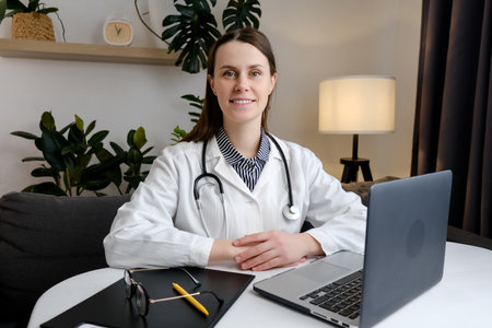 Portrait of smiling cute young brunette woman nurse sitting at desk in hospital or private clinic. Happy positive caucasian female doctor in white medical uniform at workplace. healthcare conceptの写真素材
