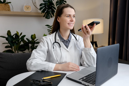 Smiling beautiful young woman doctor recording audio message on smartphone sitting at table with laptop, chatting with patient online, giving consultation or advice, treatment, using virtual voice appの写真素材