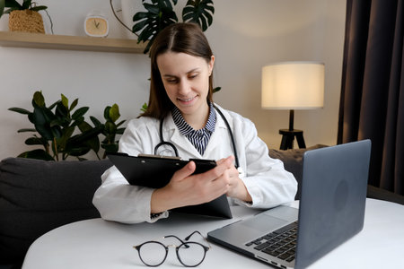 Close up of smiling doctor woman filling paper medical records. Practitioner in white coat doing paperwork at workplace with laptop, writing notes, preparing documents, reports. online doctor conceptの写真素材
