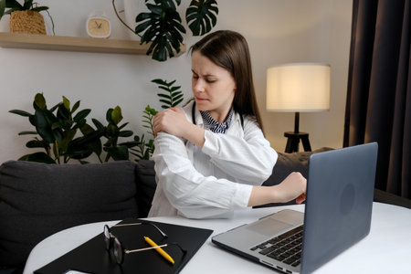 Tired woman doctor in uniform touch stiff shoulder feeling hurt joint back pain rubbing massaging tensed muscles suffer from fibromyalgia ache after long computer work study in incorrect postureの写真素材