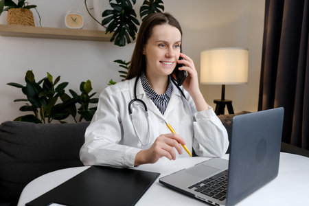 Smiling cute young female doctor physician holding cell phone talking on mobile, using laptop. Healthcare professional answering call giving remote consultation on smartphone making appointment notesの写真素材