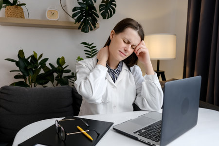Tired sad female doctor at computer experiences pain and discomfort in the neck. Concentrated woman general practitioner in office working at laptop typing on keyboard takes break and touching shoulderの写真素材