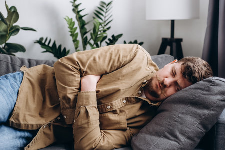 Sad young man lying on gray couch at home, suddenly feeling strong stomach ache, gastritis problem. Upset male squeezing belly with hands because of abdominal pain. Guy suffering from stomach acheの写真素材