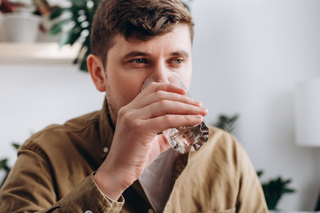Selective focus of young caucasian man drinking fresh water from glass, sit on couch in room at home. Health care, proper nutrition at home, thirsty in the morning and domestic life, hydration conceptの写真素材