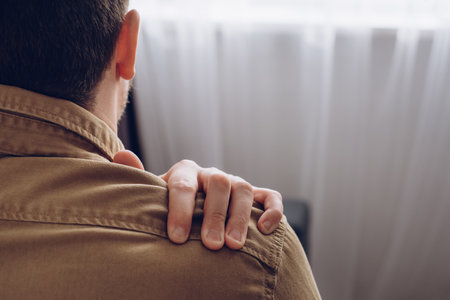 Side view of unhealthy young man sitting near window suffering from pain in shoulder, doing self-massage. neck pain. Forearm pain, people with body muscle problems, healthcare and medicine conceptの写真素材