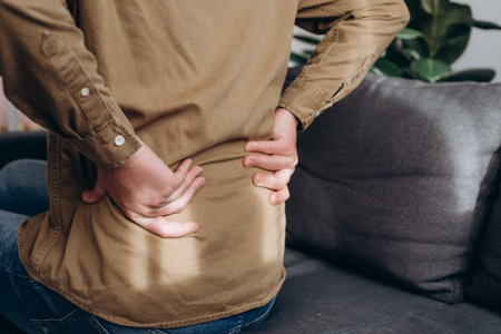 Side view of unhealthy young caucasian man sitting on couch at home suffering from back pain, have backache, health problems, lower back, has chronic joint problems. Problems with spine conceptの写真素材