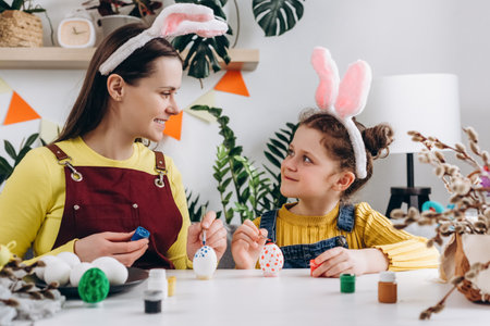 Happy young mother with smiling little kid daughter to dye and decorate eggs with paints for Easter holidays while sitting together at white table in room. Easter Family traditions and spring conceptの写真素材