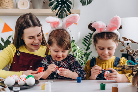 Concept of creativity and spring holiday. Happy young mother hugging two pretty little daughters, positive family painting eggs sit at table, wears pink bunny ears. Easter celebration and traditionsの写真素材