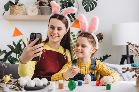 Pretty little daughter and happy young mother in fluffy bunny ears holding video call with relatives or friends using smartphone app, congratulating with Easter online, distant festive communicationの写真素材