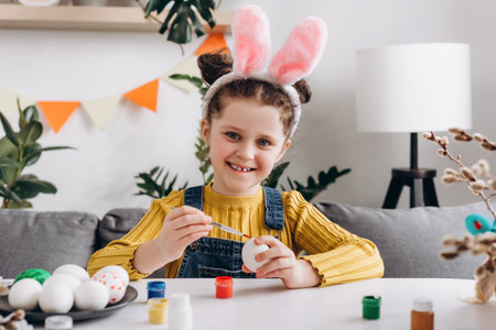 Portrait of beautiful little girl child in pink fluffy bunny ears with painting eggs sit at table in living room at home. Happy family preparing for Easter. Cute small kid positive looking at cameraの写真素材