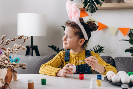 Charming dreamful little girl kid wearing fluffy bunny ears sitting at white table and painting eggs while preparing for Easter celebration, happy cute child looking at window. spring holiday conceptの写真素材