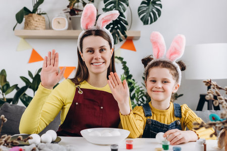 Funny young mom and little girl blogger waving hand, looking at camera, greeting, saying hello. Pretty little daughter and smiling mom in bunny ears talk on video call sitting at table. easter conceptの写真素材