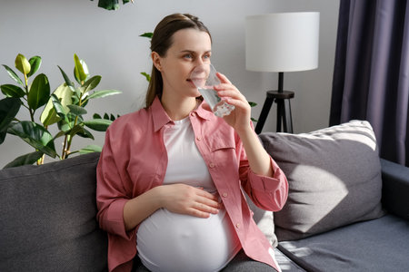 Happy peaceful young pregnant woman drinking water sitting on cozy sofa at home. Beautiful expectant lady have rest in living room with glass, stroking big belly. Pregnancy, healthcare, thirst conceptの写真素材
