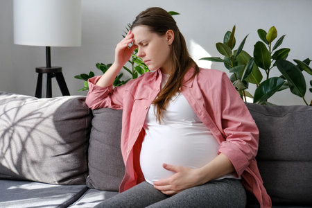 Worried young pregnant female feeling stress and anxiety while sitting alone on couch in living room at home. Expectant mother thinking about the upcoming birth of child. depression and motherhood conceptの写真素材