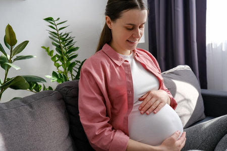 happy maternity concept. Smiling young single pregnant woman sitting on cozy couch in light room touch big belly enjoy late stages of pregnancy. Caring mother strokes pregnant tummy with two handsの写真素材