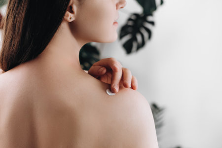 Close up back view of young female 20s old years applying cream on shoulder standing in cozy light bathroom at home. Beautiful lady apply body lotion. Health and beauty concept. Daily skincare routineの写真素材