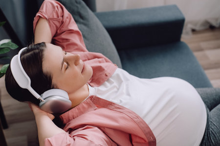 Relaxed attractive young pregnant woman listening to music with wireless headphones resting on cozy sofa in living room at home. Beautiful happy future mother, modern technology and interiors conceptの写真素材