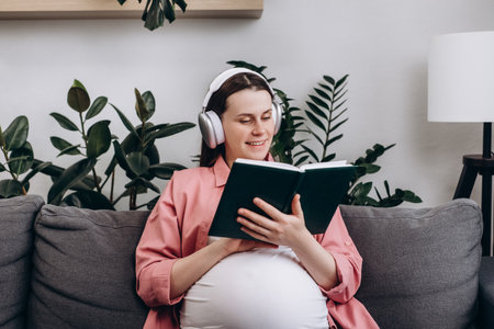 Smiling young Caucasian pregnant woman wear wireless headphone listen relax music and reading book to tell story to unborn child. Happy calm future mother sitting on couch in living room at homeの写真素材