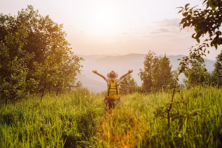 Happy traveler little girl in hat and yellow backpack raises his hands up on background amazing mountains. Child enjoying beautiful nature at warm sunset or sunrise. freedom and imagination conceptの写真素材