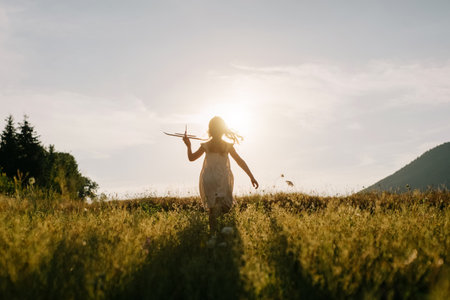 Silhouette of preteen girl kid run with airplane on background majestic mountains during warm summer sunset. Dream freedom concept. Playful child runs on green meadow holding in hands toy aircraftの写真素材