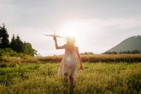 Little girl holding airplane toy during running in green field during warm summer day. Dream freedom concept. Playful cute little child pilot on background mountain and sky at amazing sunsetの写真素材