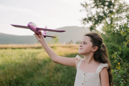 Portrait of happy teen girl in dress playing with violet toy airplane during amazing warm sunset or sunrise. Childhood dreams. Family, nature, freedom and airplane concept. Playful cute child pilotの写真素材
