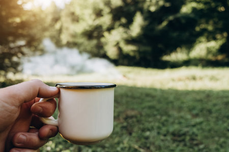 Close up of unknown young caucasian man holding hot coffee near beautiful bonfire in summer morning forest. Hiking guy enjoying warm nature. Wanderlust adventures in outdoor living lifestyle, hikingの写真素材