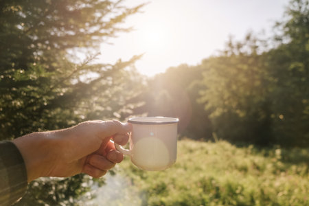 Close-up of male hand holding cup of coffee on background forest and sky during warm sunrise or sunset. Tourist male drink hot tea and enjoy scenery in nature. Trekking and wanderlust conceptの写真素材