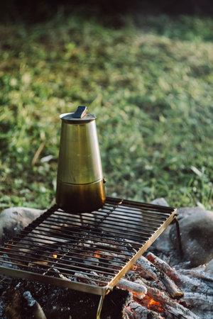 Close-up of geyser coffee maker over warm bonfire in green summer park during sunrise or sunset. Camping vibes, nature and outdoor lifestyle mood concept. Campfire in summertime, hot coffeeの写真素材