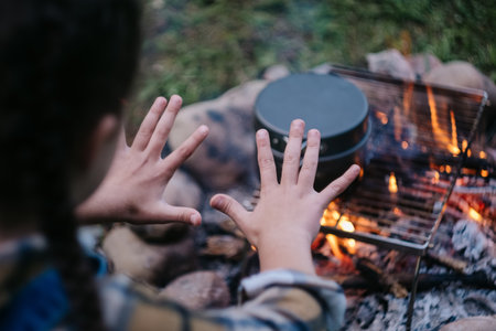 Close up of unrecognizable little girl child warming up hands next to warm calm campfire at evening time. Hiking, adventure active vacations, travel, camping vibes and outdoor lifestyle moodの写真素材