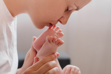 Happy caring young caucasian mother playing with baby at home, smiling loving mom kissing toddlers feet sitting on cozy couch, having fun parent bonding with child enjoying motherhood childcareの写真素材
