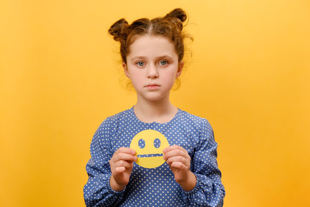 Portrait of unhappy upset preteen caucasian girl child holding sad yellow emoticon, looking at camera, posing isolated over plain yellow color background wall in studio. Feedback rating conceptの写真素材