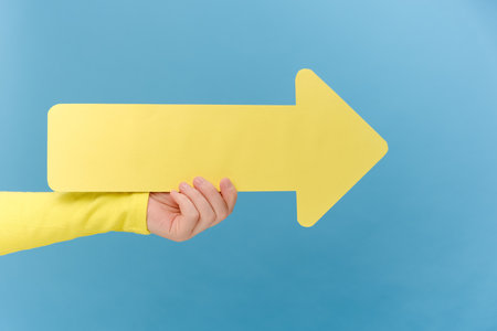 Close up of woman hand holding big yellow arrow showing direction, indicating sign, posing isolated over plain blue color background wall in studio with copy space for advertisement or designの写真素材
