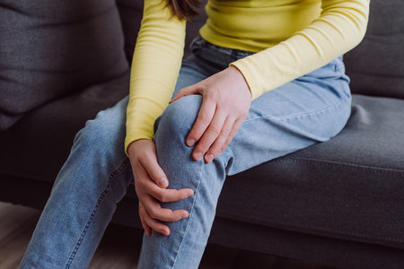 Close up of unknown young caucasian female touching her knee feel pain and doing massage sitting alone on sofa at home. People health care bone problem, illness, cramps, rheumatism conceptの写真素材
