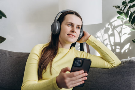 Relaxed pretty brunette girl wearing earphones listening to mobile music playing via smartphone app,...