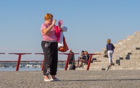 DNEPR, UKRAINE - OCTOBER 02, 2016:Mature woman talking on cellular using speaker standing on a Dnepr river embankment in center of the Dnepr city at warm and sunny autumnal weekendのeditorial素材