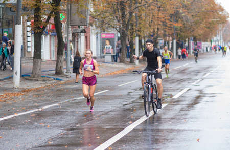 DNIPRO, UKRAINE - SEPTEMBER 16, 2018: Young man on a bicycle shooting  video of young woman running  during 21 km distance of ATB Dnipro Marathon on rainy Dnipro city streetのeditorial素材