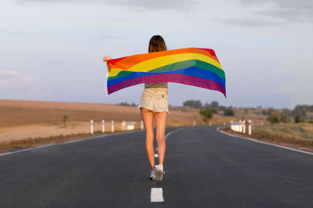 Young european lesbian woman with lgbt rainbow flag at the road at sunset. Fit woman with brown hair in a denim shorts posing with lgbt pride flag.の写真素材