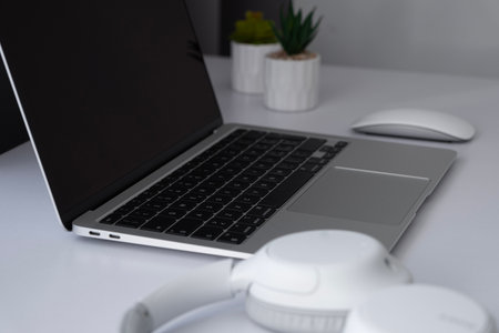 Side view on a blank screen laptop with mouse, headphones and plants on a white table at home...