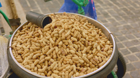 Roasted peanuts heaped in bowl, unshelled shells filling metal container, street vendor display at market, earthy brown tones, crunchy texture, protein snack ready for sale, busy outdoor stallの写真素材
