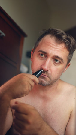 Vertical photo Man shaving at bathroom, White man carefully grooming facial hair, Man using trimmer to tidy up around nose and chin, Man in private bathroom carefully trimmingの写真素材